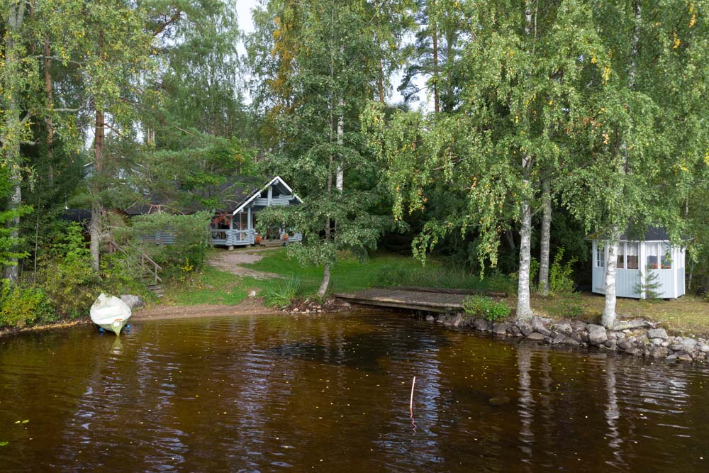 Sini-lomamökki järven rannalla, laiturin vieressä soutuvene ja rantasauna puiden suojassa.