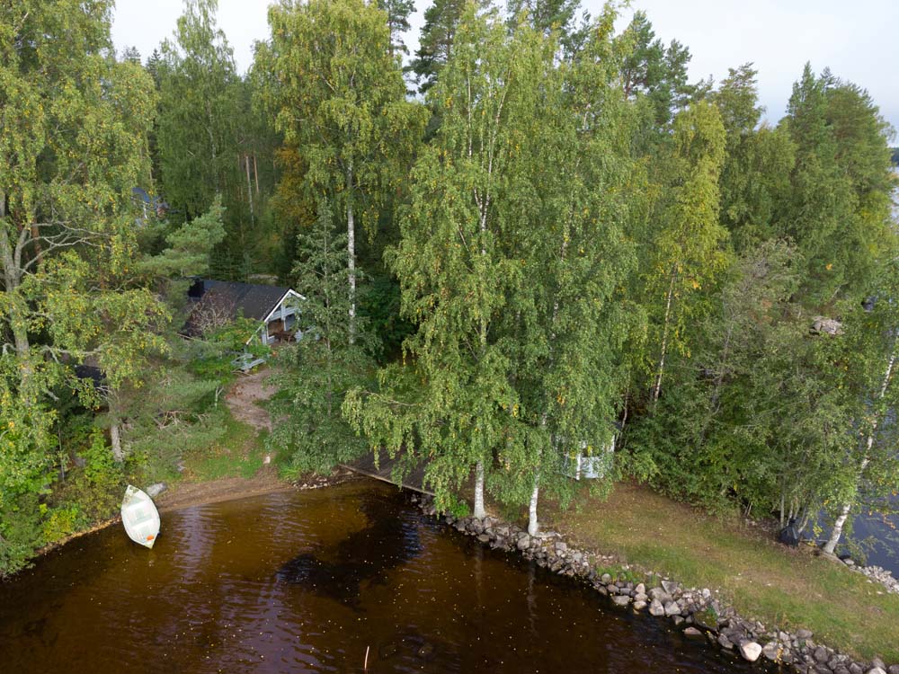 Hiekkarannan mökki näkyy puiden keskellä Kannonjärven rannalla, ja rannassa on soutuvene sekä kivinen pengerrys.