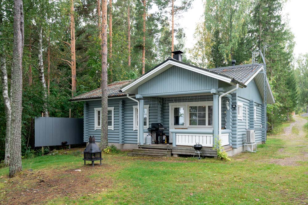 Kulmala-lomamökki metsän keskellä, pihalla terassi, pallogrilli ja tulisija.