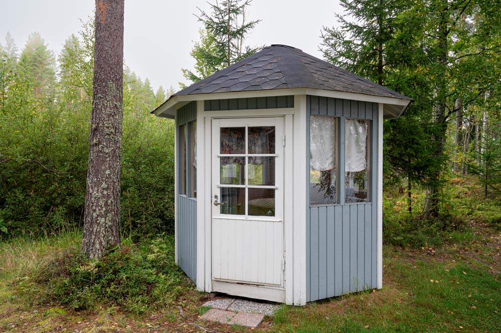 Pieni kahdeksankulmainen puinen piharakennus metsäisellä pihalla.