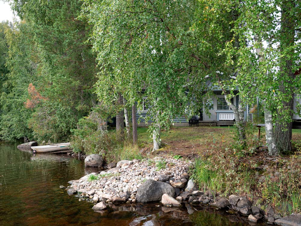 Hirsimökki järven rannalla puiden katveessa, pienen laituriin kiinnitetyn soutuveneen vieressä.