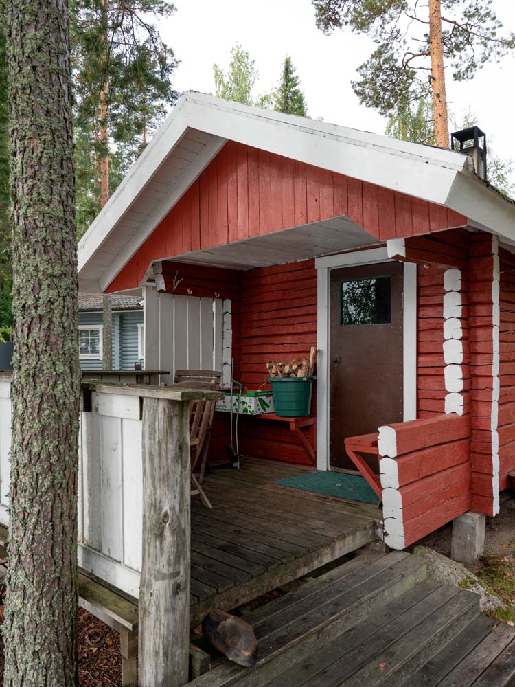 Punainen lomamökin sisäänkäynti puisine kaiteineen ja portaineen mäntypuiden keskellä.