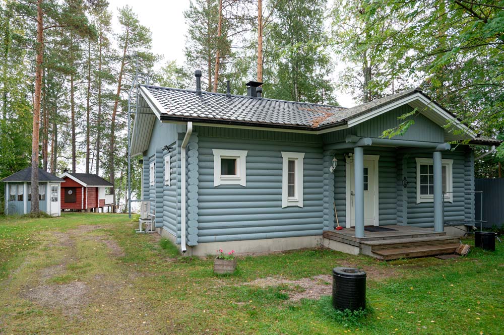 Harmaa hirsimökki ja pihapiiri metsäisessä ympäristössä, taustalla pieni piharakennus.