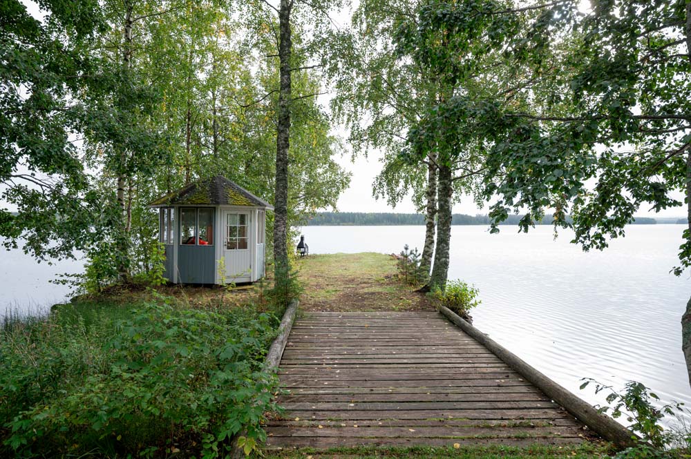 Pieni rantasauna ja puinen laituri järven rannalla koivujen keskellä.
