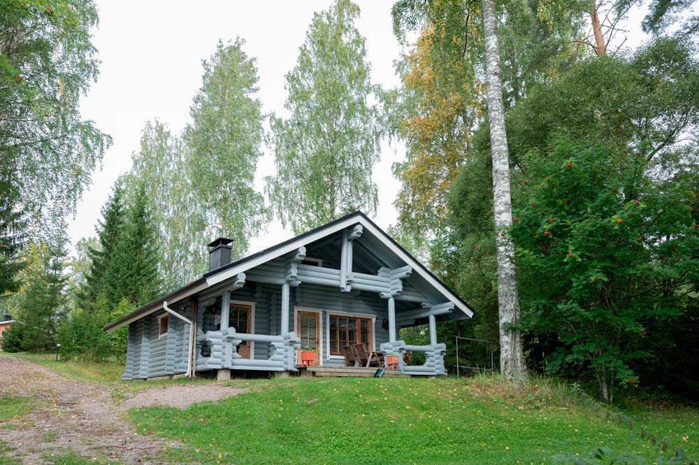 Sini-lomamökki metsän reunassa, edessä nurmipiha ja terassi.