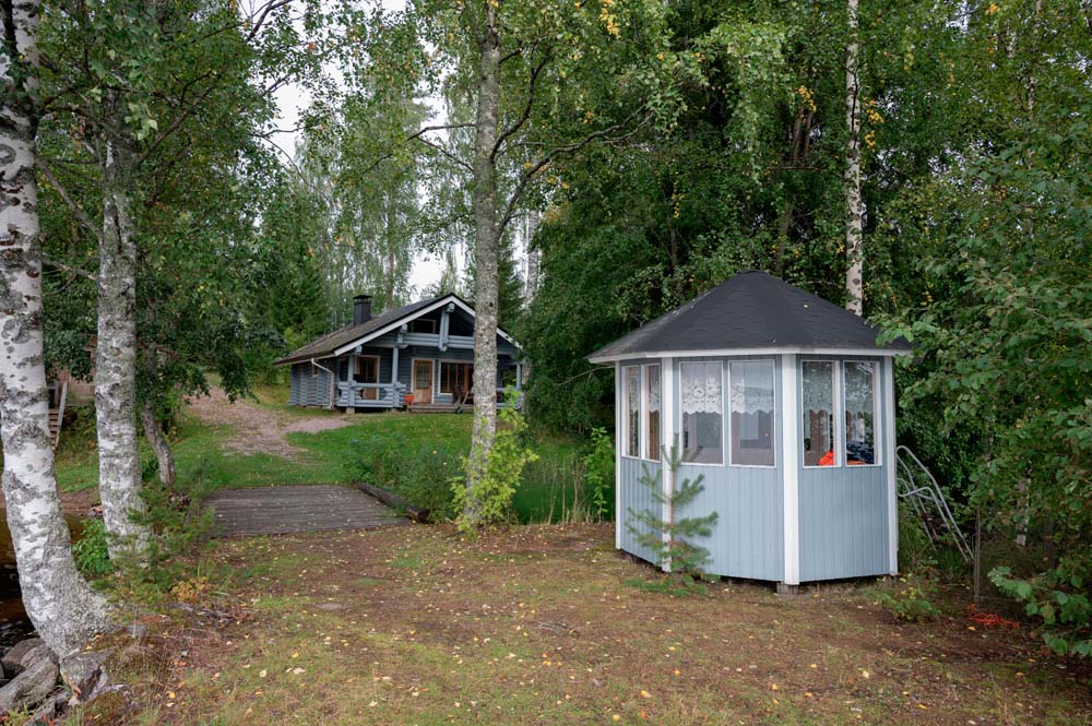 Sini mökki, Kannonkoski Pihalla on pieni kuusikulmainen huvimaja, ja taustalla näkyy siniharmaa lomamökki puiden keskellä.