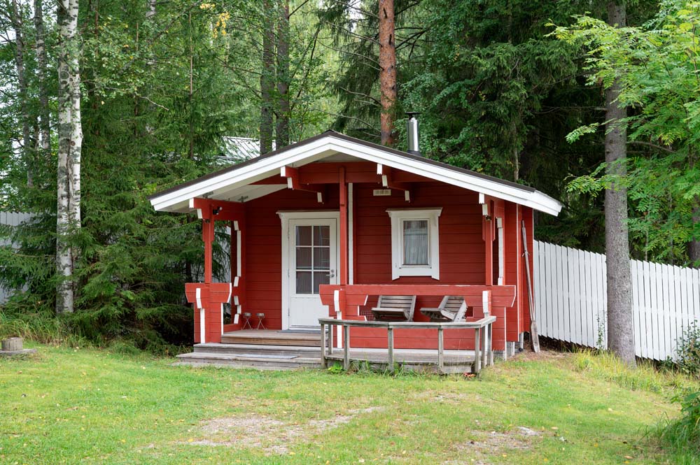 Punainen rantasauna metsän reunassa, edessä pieni terassi ja penkit.